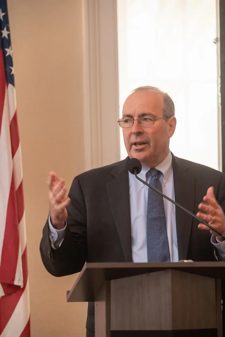 Man in dark suit and glasses gestures while speaking at a podium beside an American flag