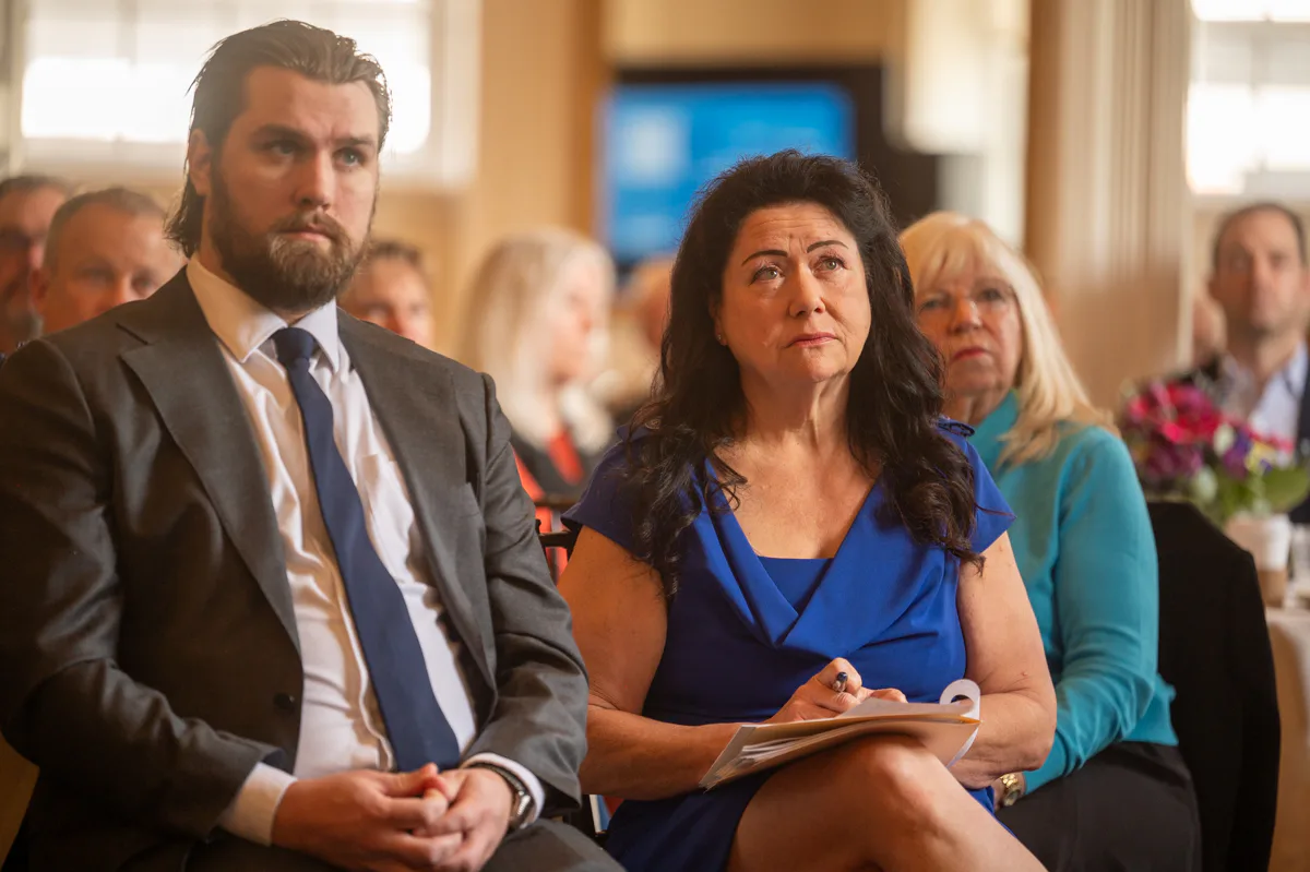 Bearded man in suit beside a woman in blue, taking notes at a seated event