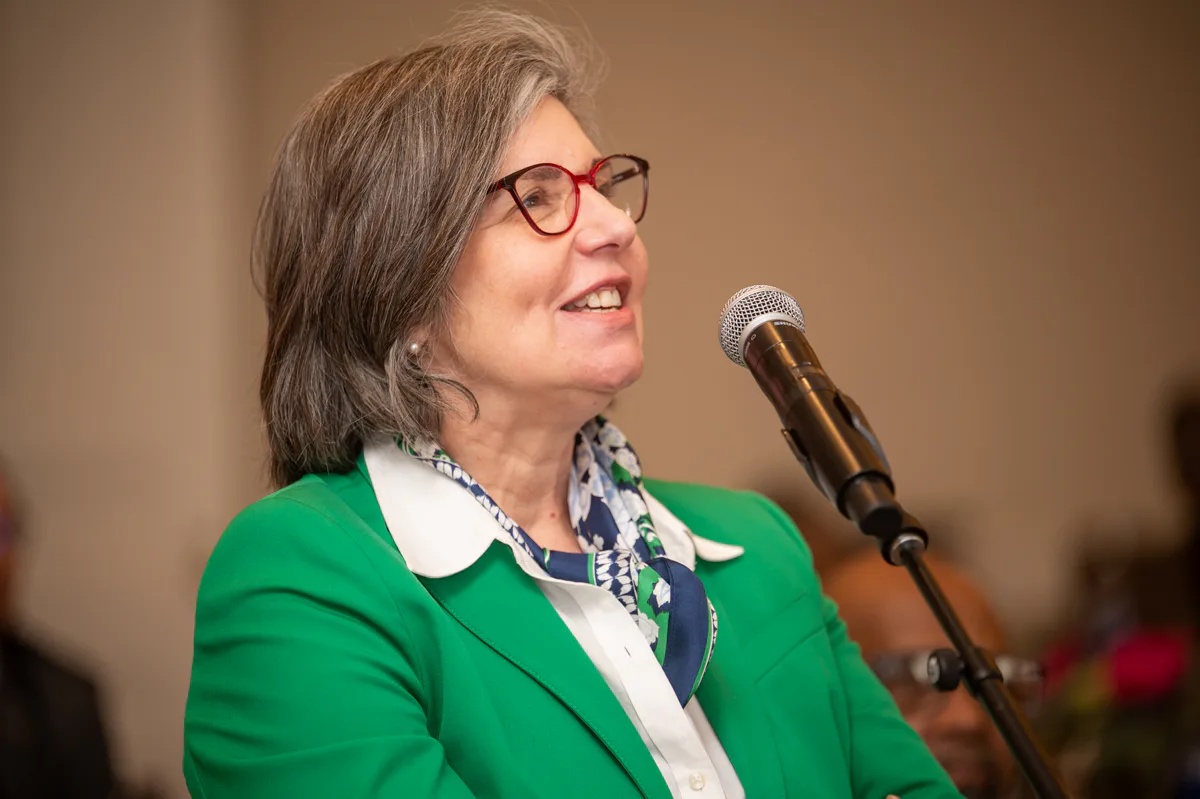 Woman with red glasses and green blazer speaks into a microphone at an event