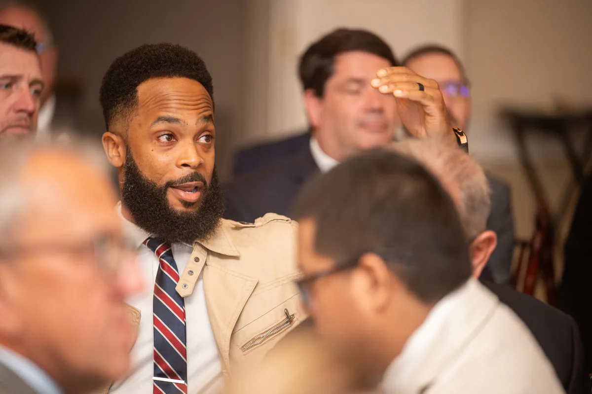 Bearded man in tan jacket and striped tie speaks with raised hand among seated attendees
