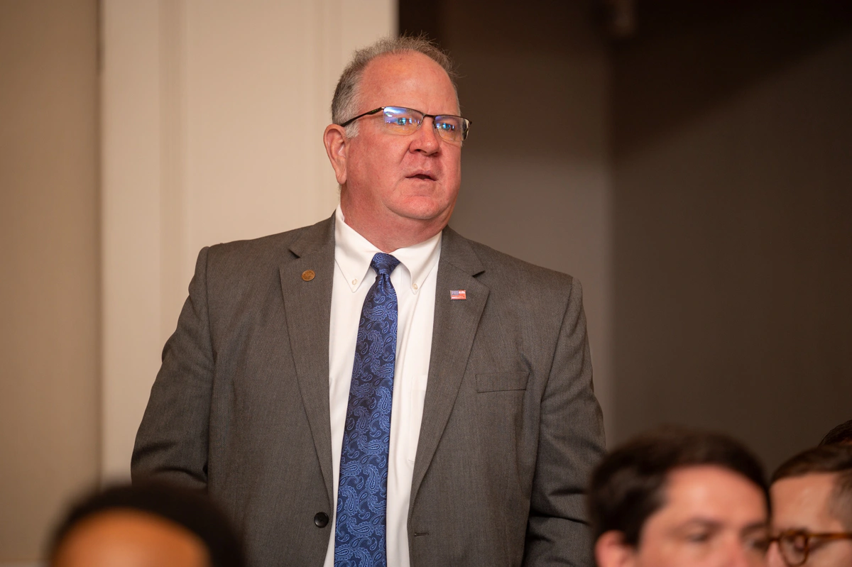 Man in gray suit and glasses stands in a crowd, looking toward the front of the room