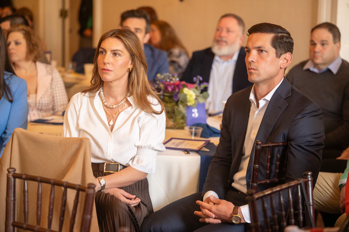 Woman in white blouse and man in dark suit sit at an event with an audience behind them