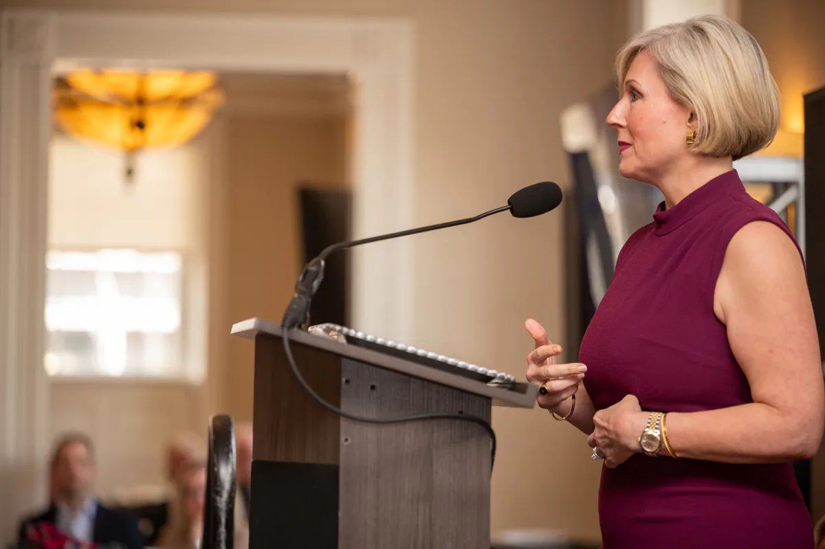 Woman with short blonde hair in maroon dress speaks at a podium beside a chandelier