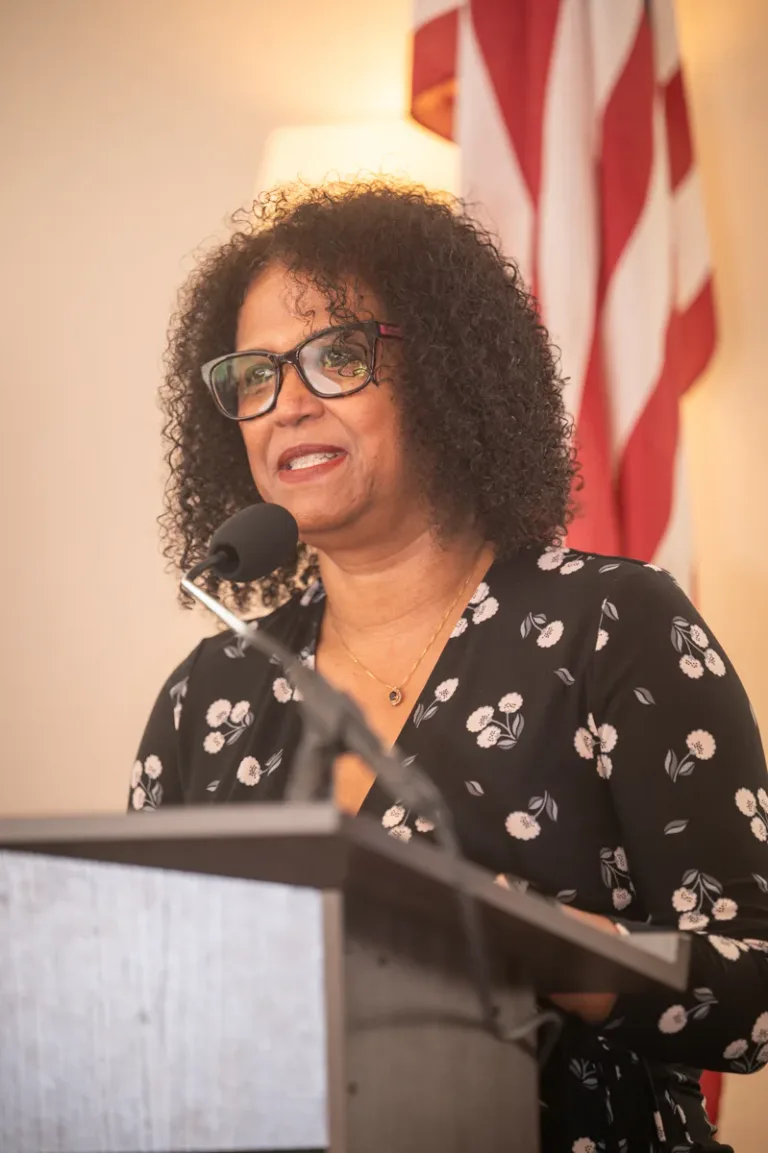 Woman with glasses and black floral blouse speaks at a podium in front of an American flag
