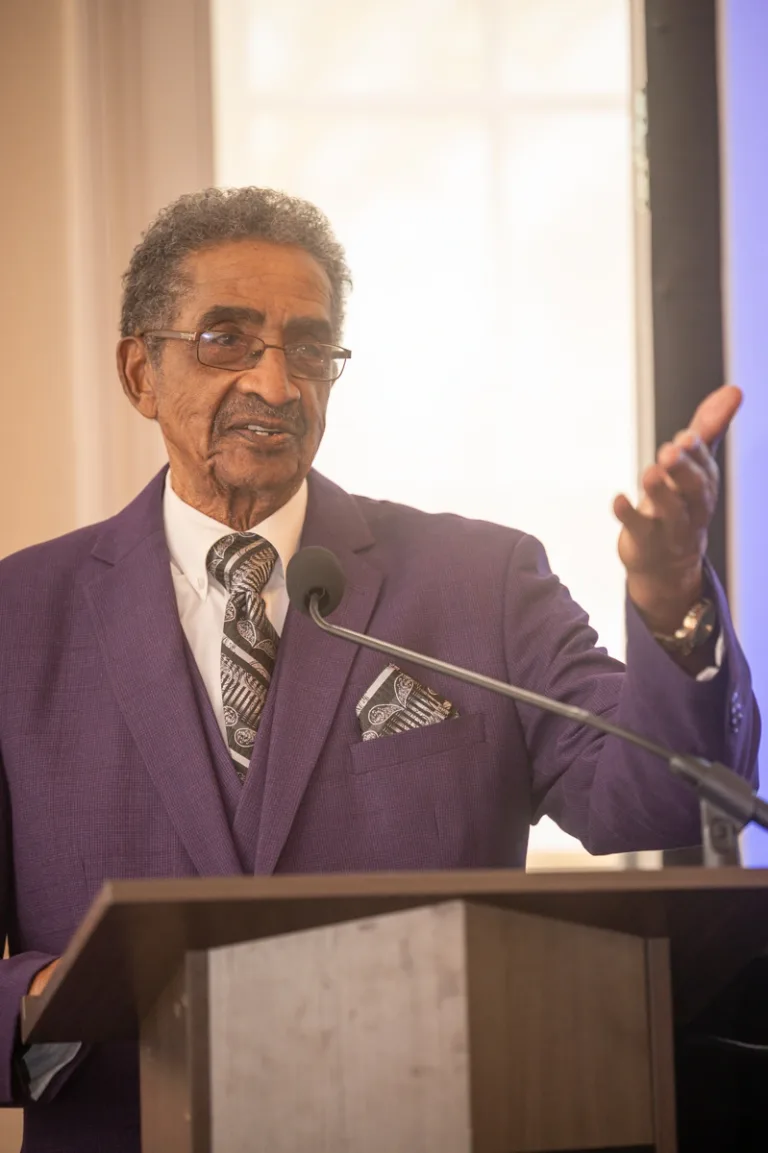 Older man in purple suit gestures while speaking at a podium