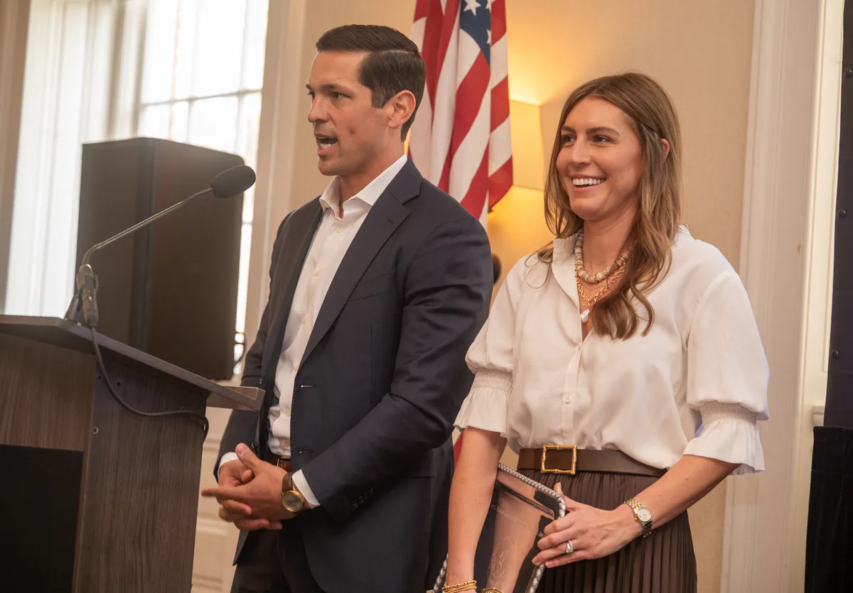 Man in dark jacket and woman in white blouse smile beside a podium with an American flag