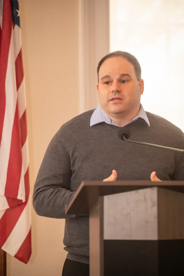 Man in gray sweater and collared shirt gestures while speaking at a podium