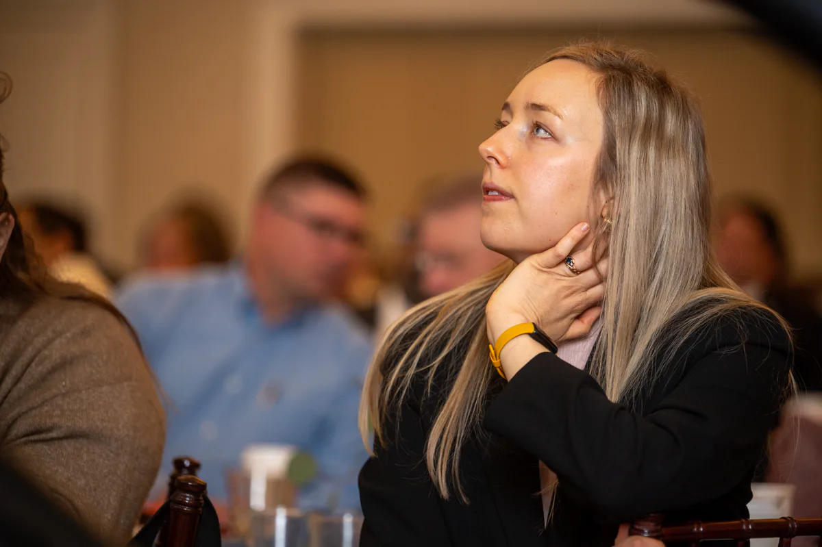 Blonde woman with long hair looks upward, hand at her neck, at a seated event