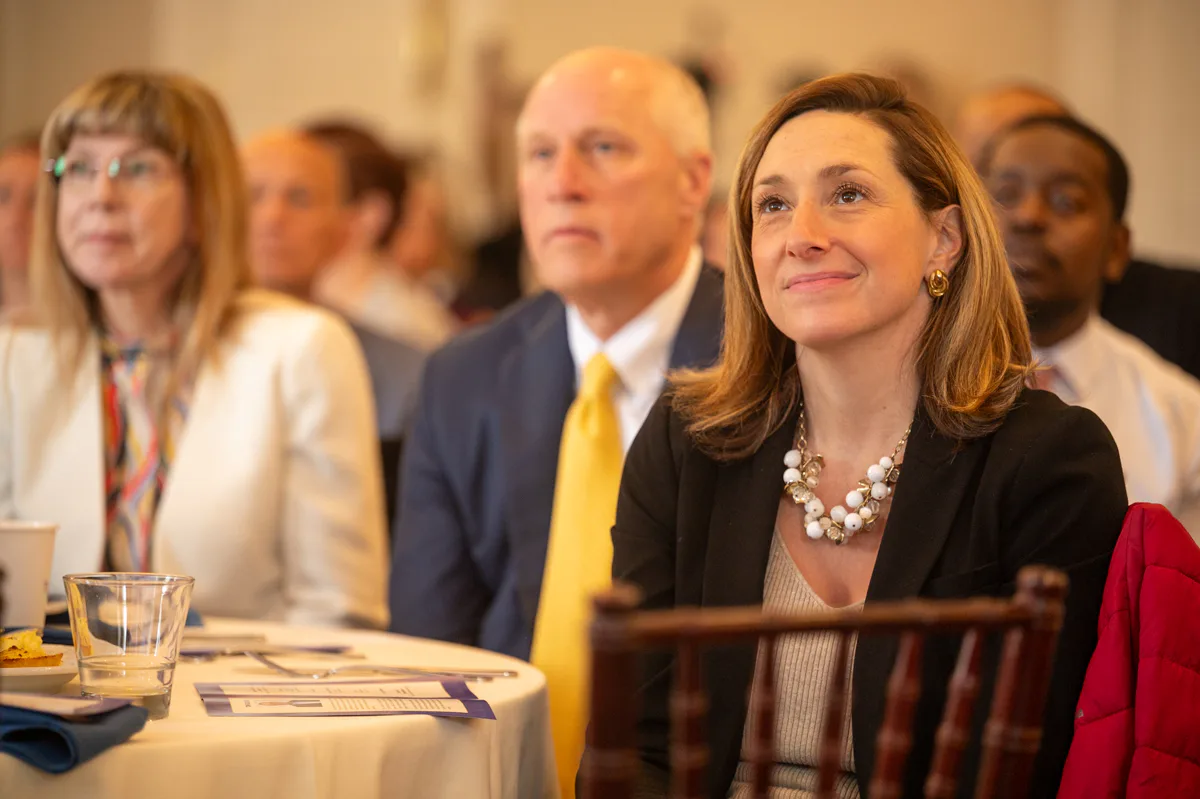Woman in pearls and older man in yellow tie sit at an event table with an audience behind