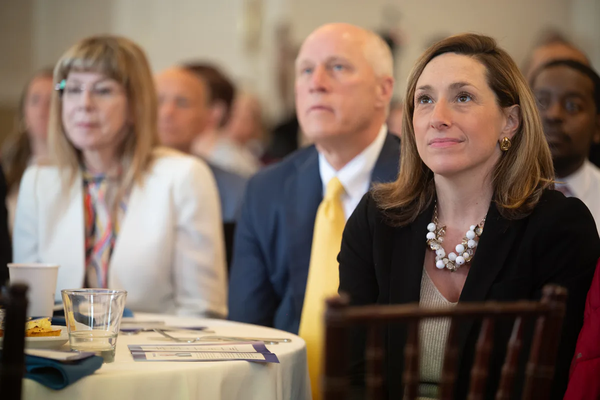 Woman in pearls and older man in yellow tie sit at an event table with an audience behind