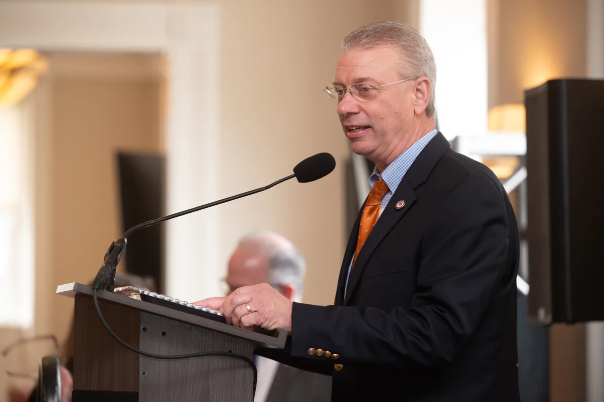 Man with glasses and orange tie speaks at a wooden podium