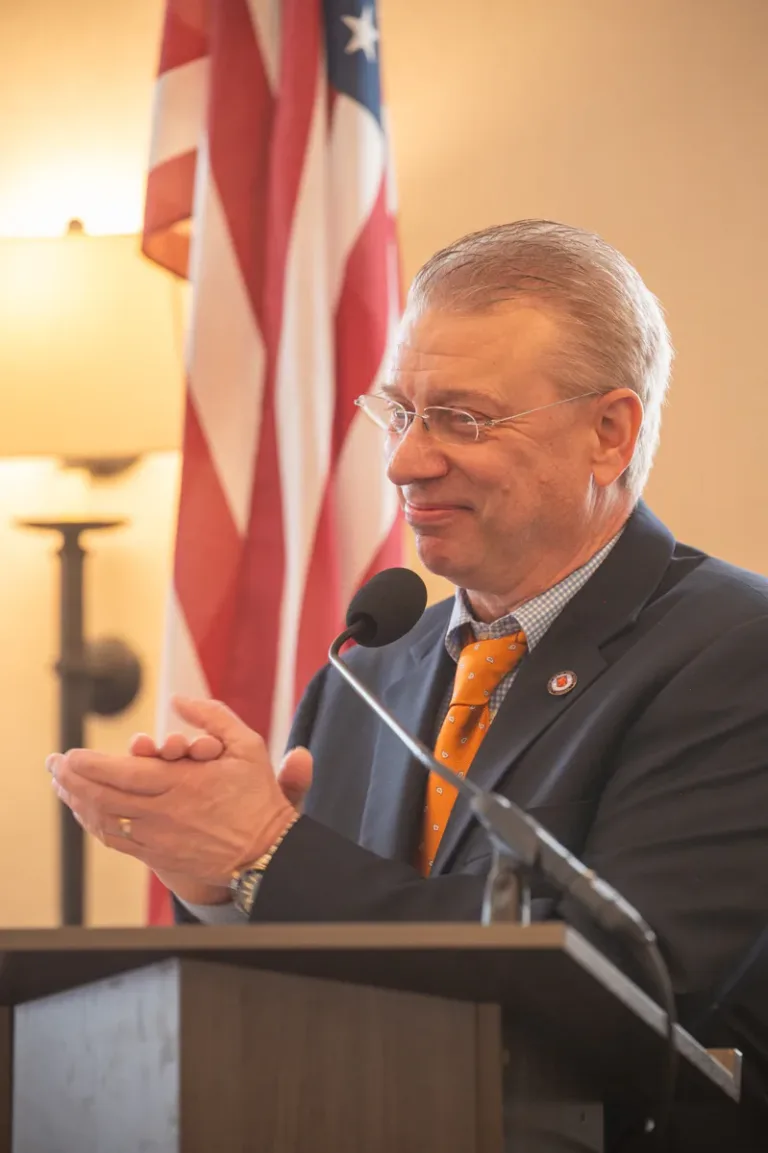 Older man with glasses smiles and claps hands together at a podium beside an American flag