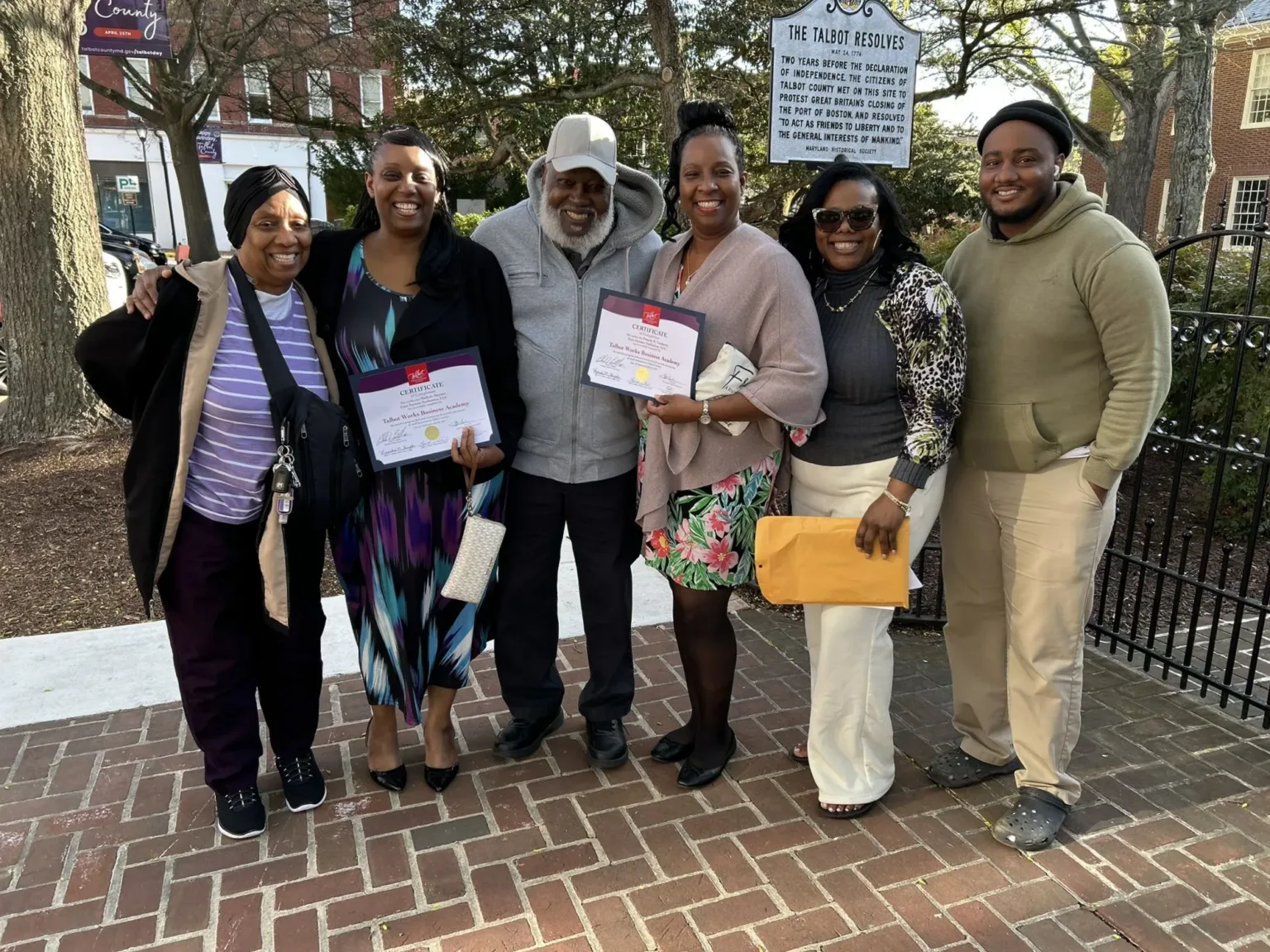 Group of six holding certificates outdoors in front of a brick building