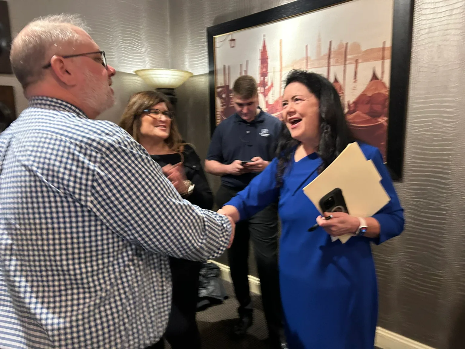 Man in checked shirt greeting a woman in a blue dress at an indoor event
