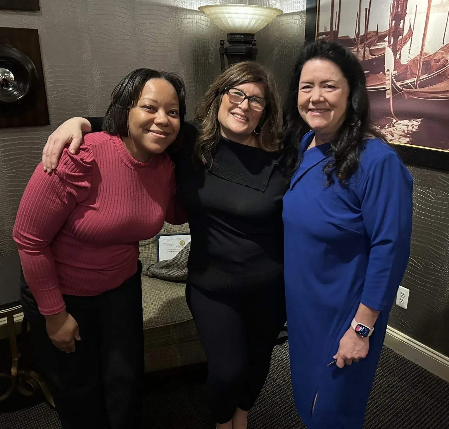 Three women posing together indoors, one in a black top and one in blue