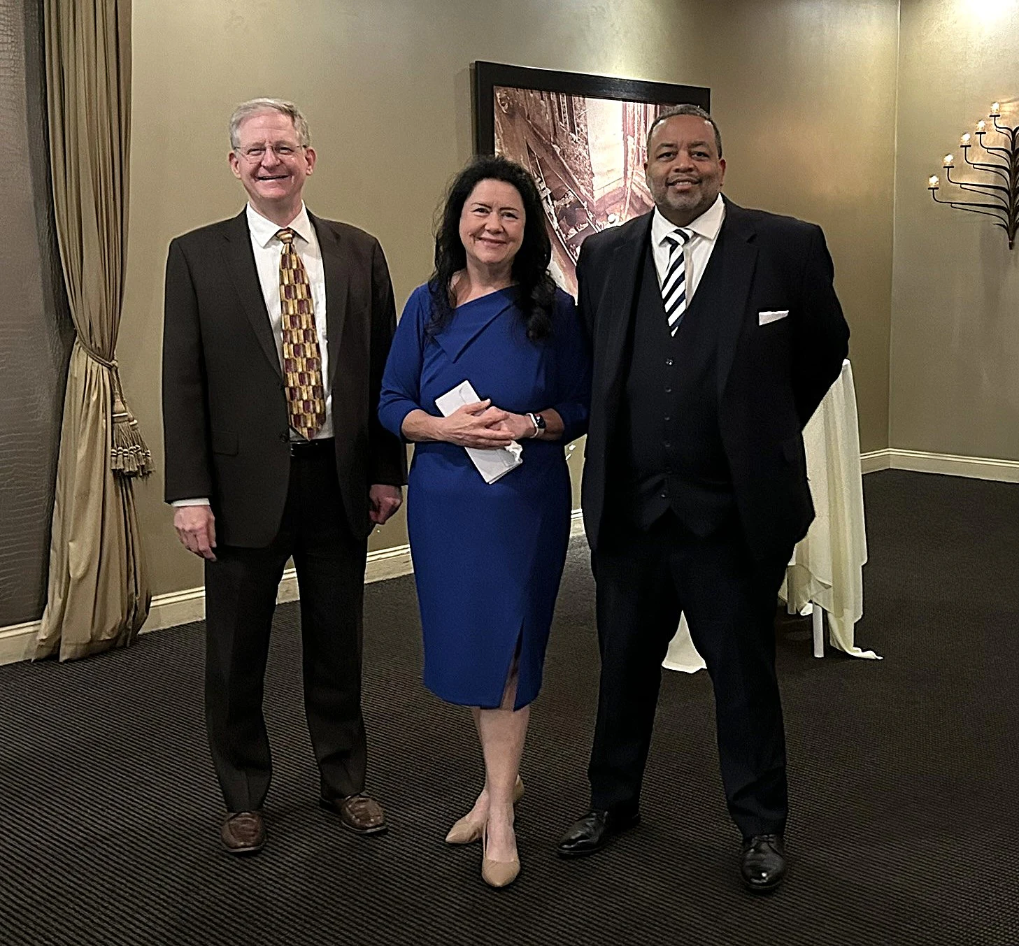 Three people posing in a room: two men in suits and a woman in a blue dress