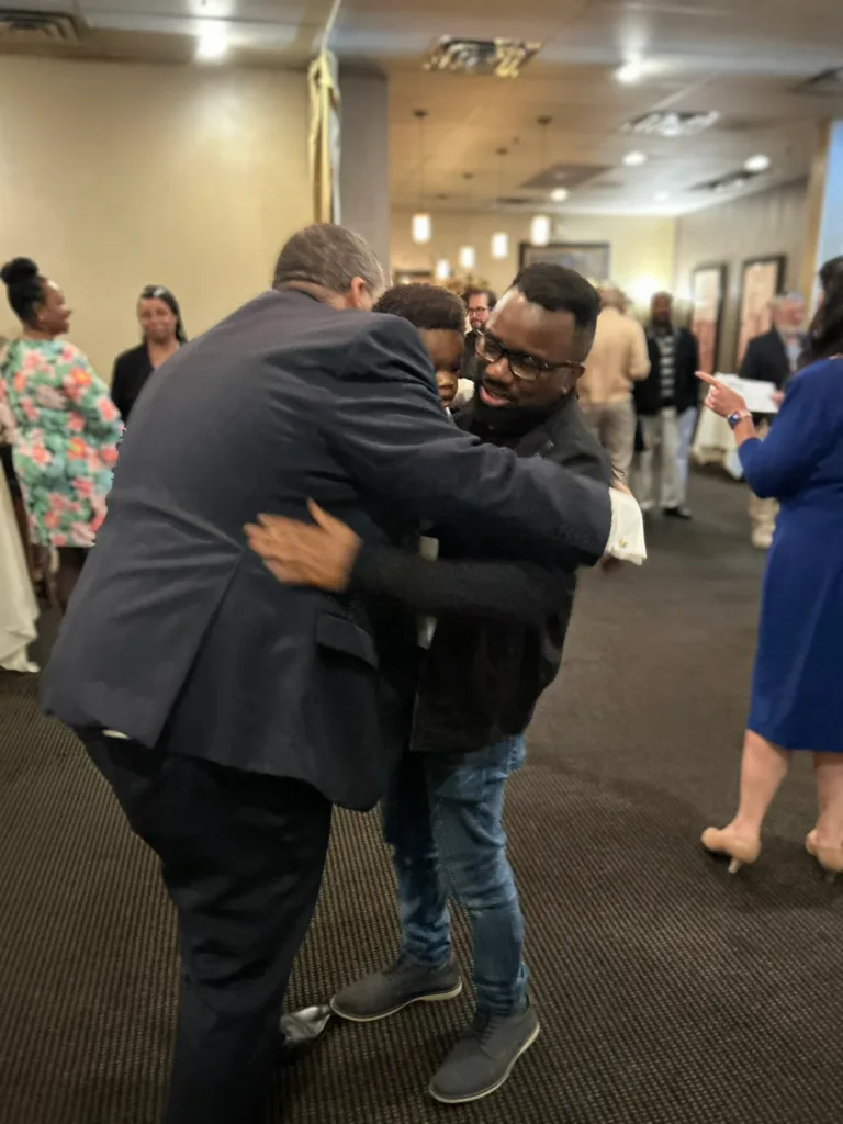 Man in dark jacket hugging a younger man in a reception room