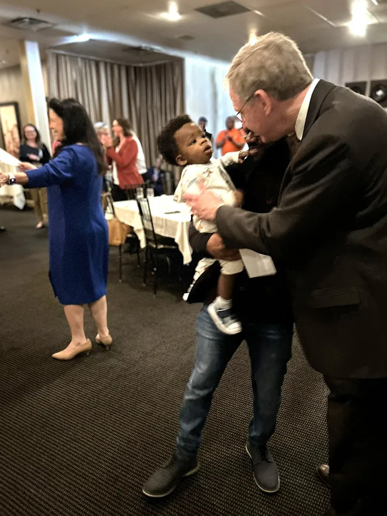 Man in dark suit bending to lift a toddler at a reception