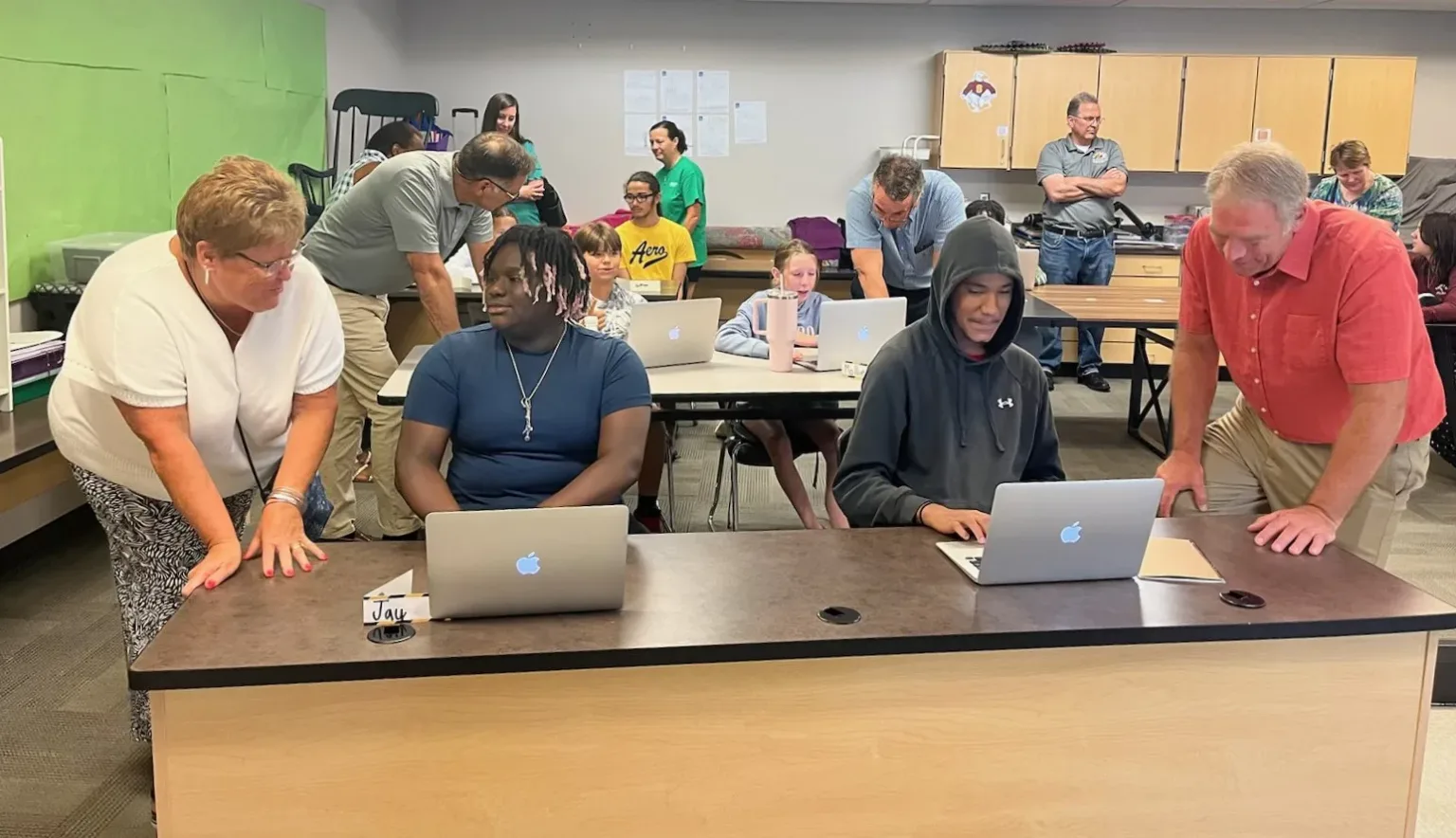 Two adults guide young people working on laptops at a classroom table.