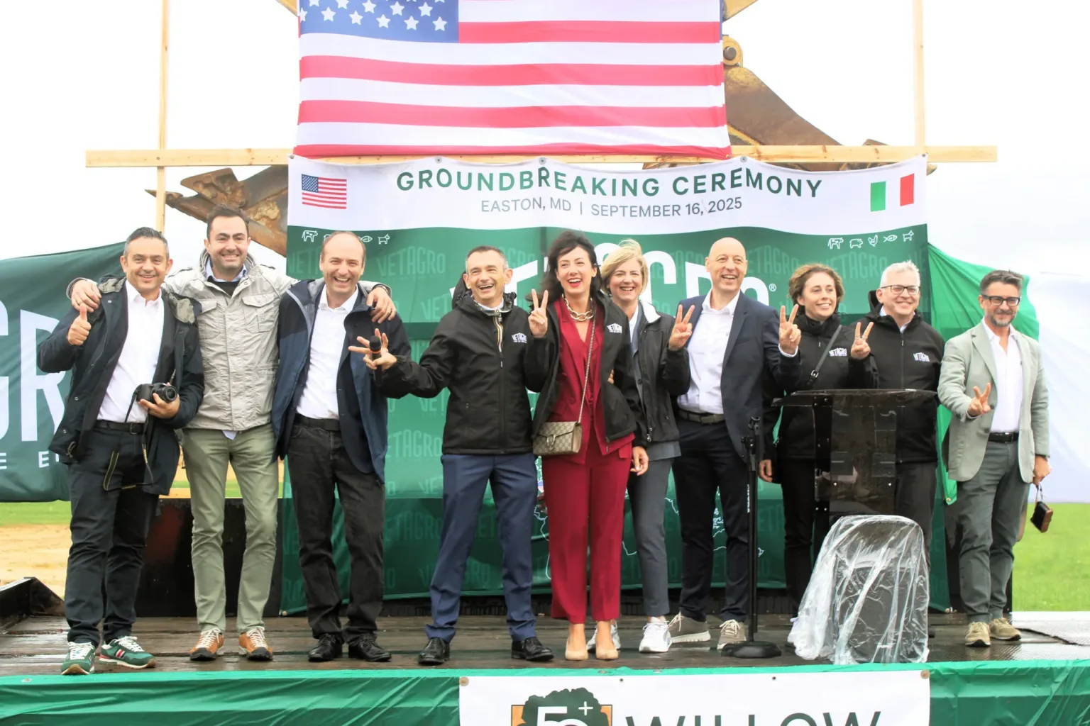 Group on a stage under a Groundbreaking Ceremony banner and American flag.
