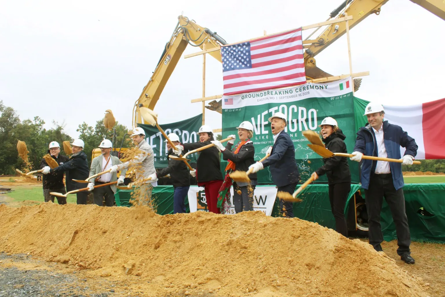 Officials in hard hats hold shovels at a groundbreaking, with a Vetagro banner and dirt pile.