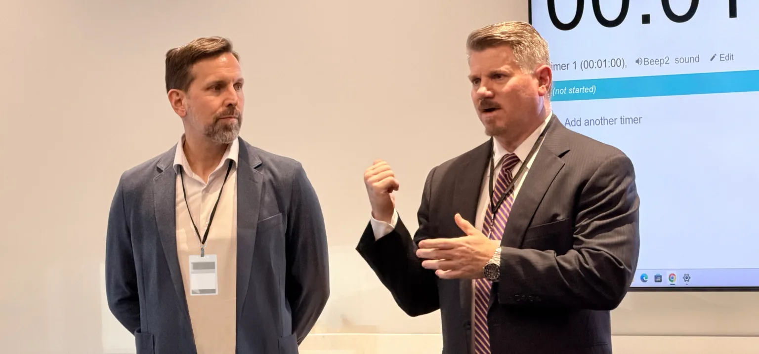 Two men in a meeting room; one gestures while presenting as the other watches.