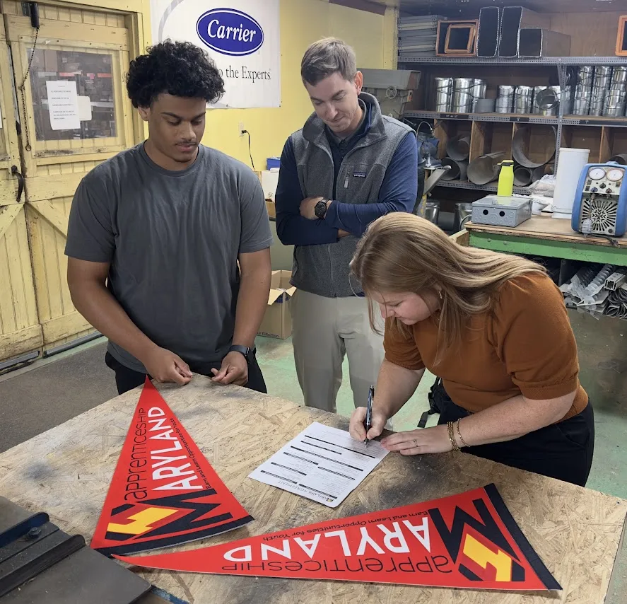 Woman signs a form at a workshop table while two men watch, with Apprenticeship Maryland pennants on the table.