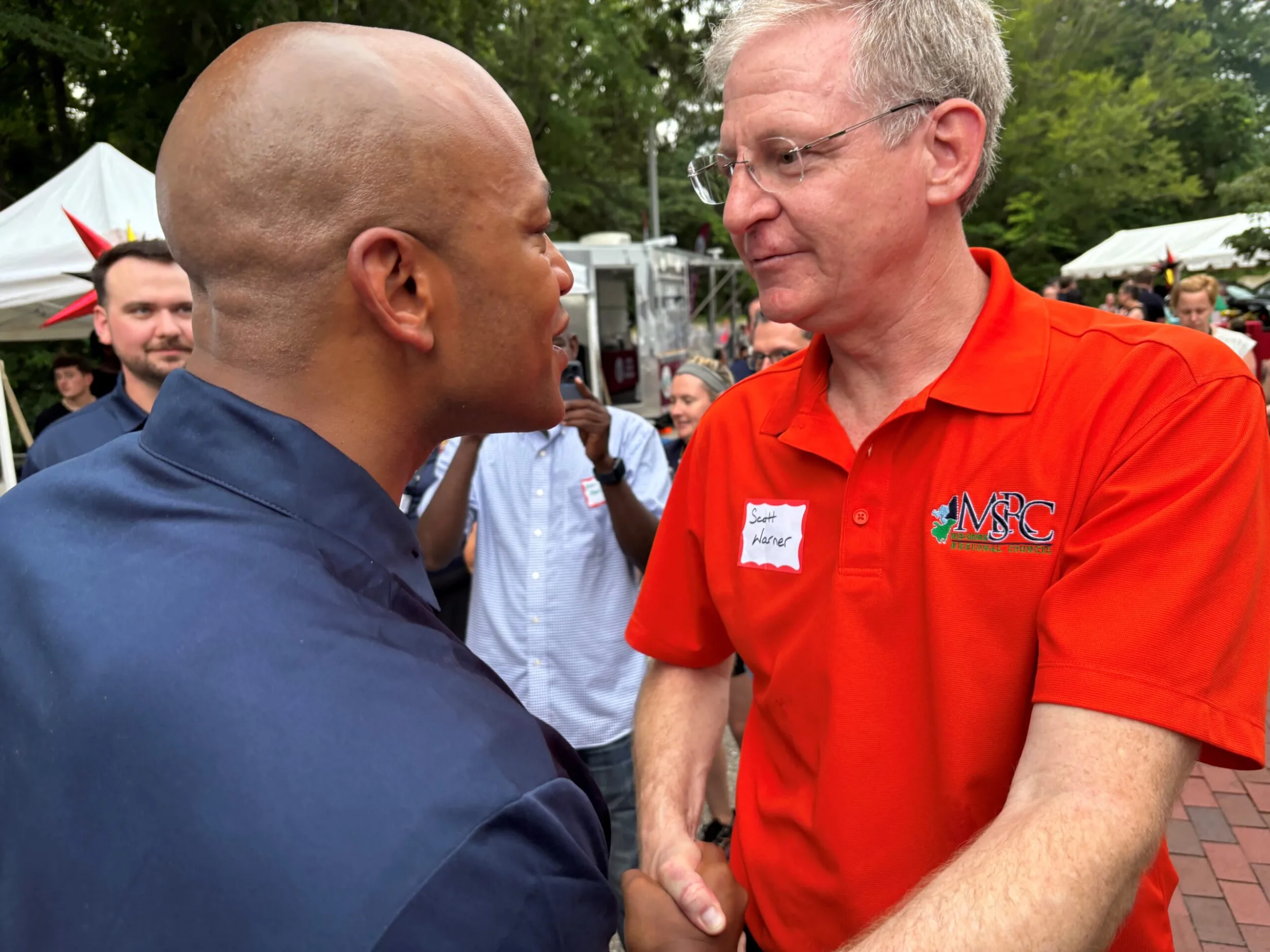 Two men shaking hands at an outdoor event, one in an orange MSRC polo