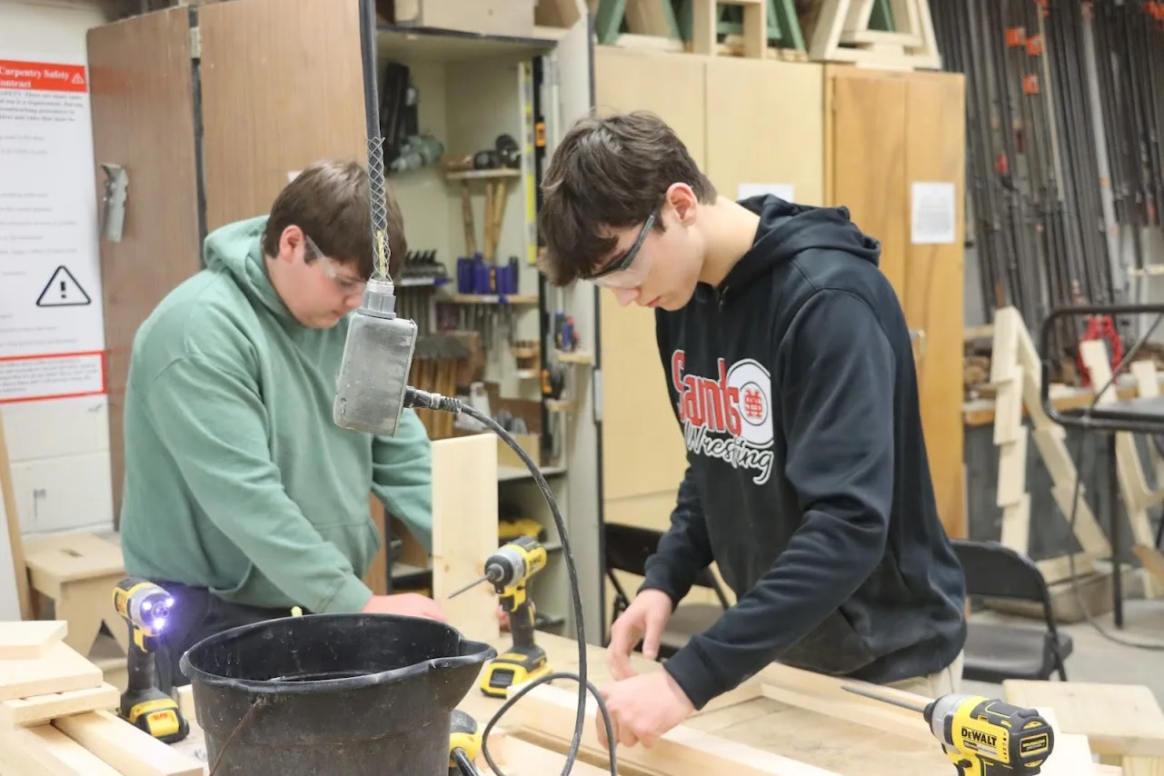 Two students in safety glasses use cordless drills on a wooden frame in a shop.