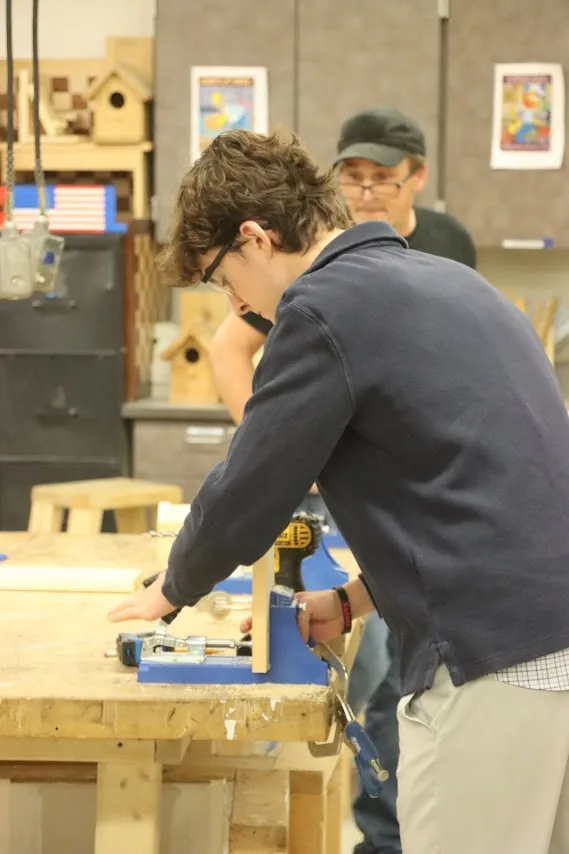 Student in a navy sweater uses a drill on a workbench, with an adult watching behind.