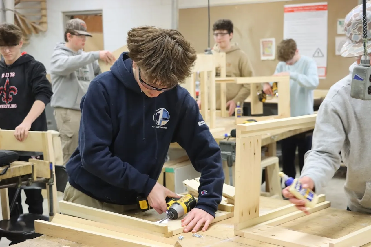 Students in safety glasses build wooden frames with drills and glue in a shop.