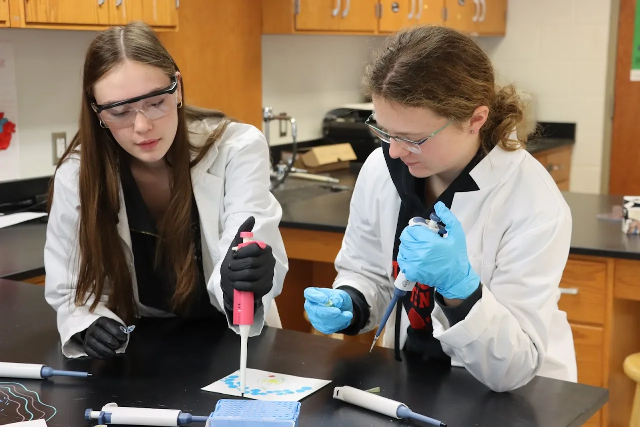 Two students in lab coats use pipettes at a lab bench.