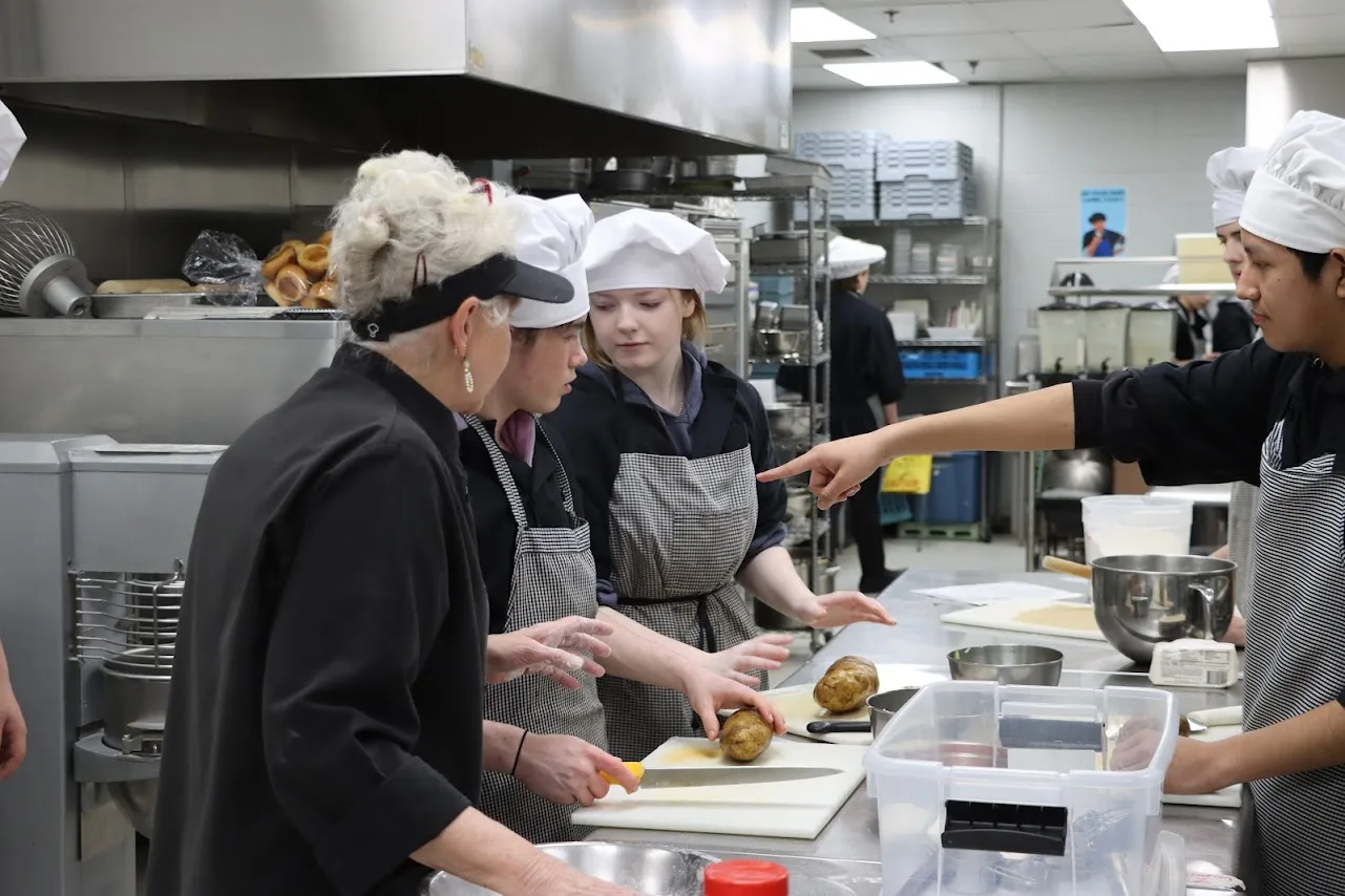 Students in chef hats and aprons prepare potatoes under an instructor's guidance in a kitchen.