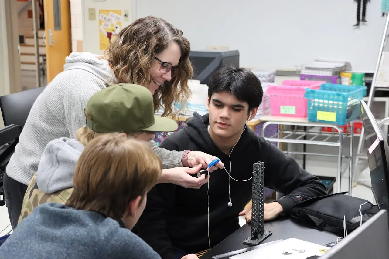 Teacher helps students with a small electronic device at a desk.