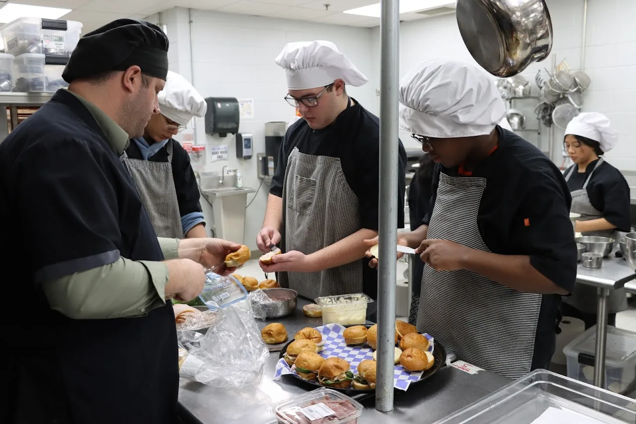 Chef instructor and students in chef gear assemble sandwiches on a metal counter.
