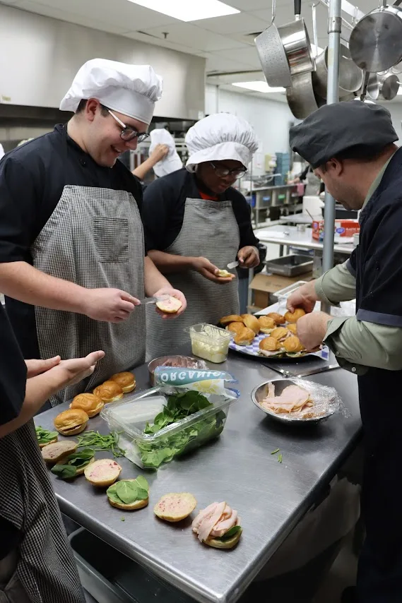 Students in chef hats and aprons build sandwiches on rolls in a commercial kitchen.