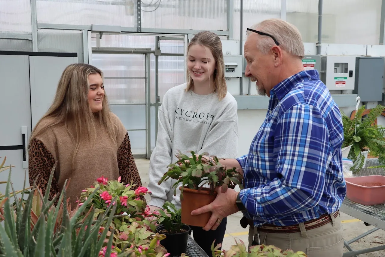 Two students and a man in plaid look at a potted plant in a greenhouse.