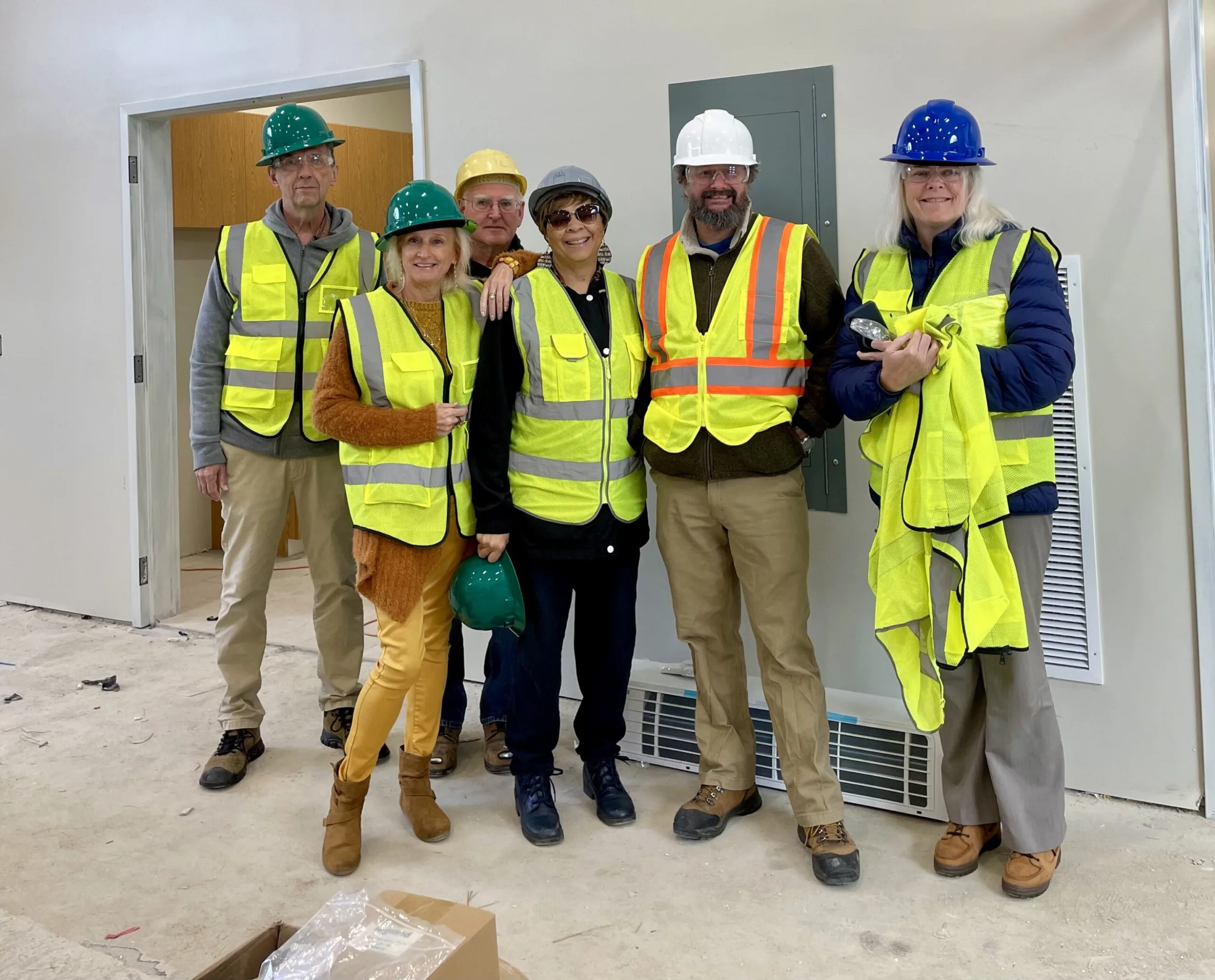 Six people in hard hats and yellow safety vests standing in an unfinished building