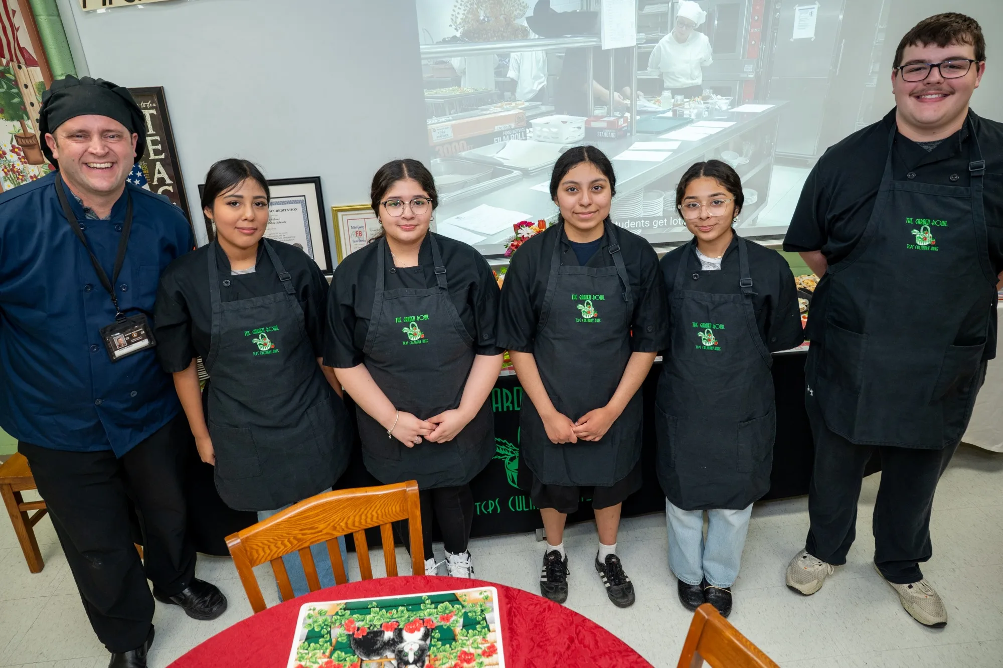 Five students and an instructor in black Garden Bowl aprons stand smiling in a classroom.