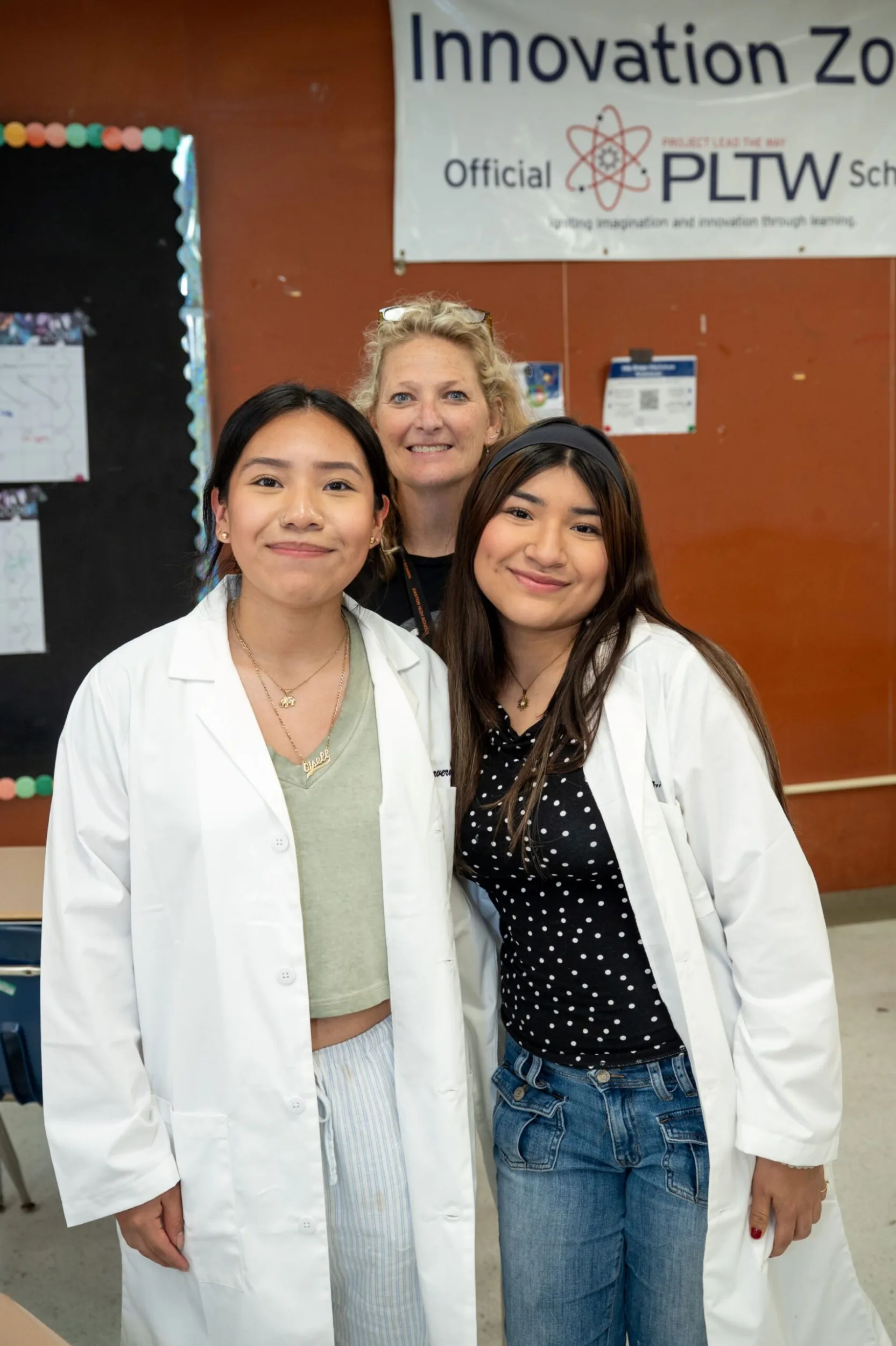 Two students in white lab coats pose with a teacher beneath an Innovation Zone PLTW banner.
