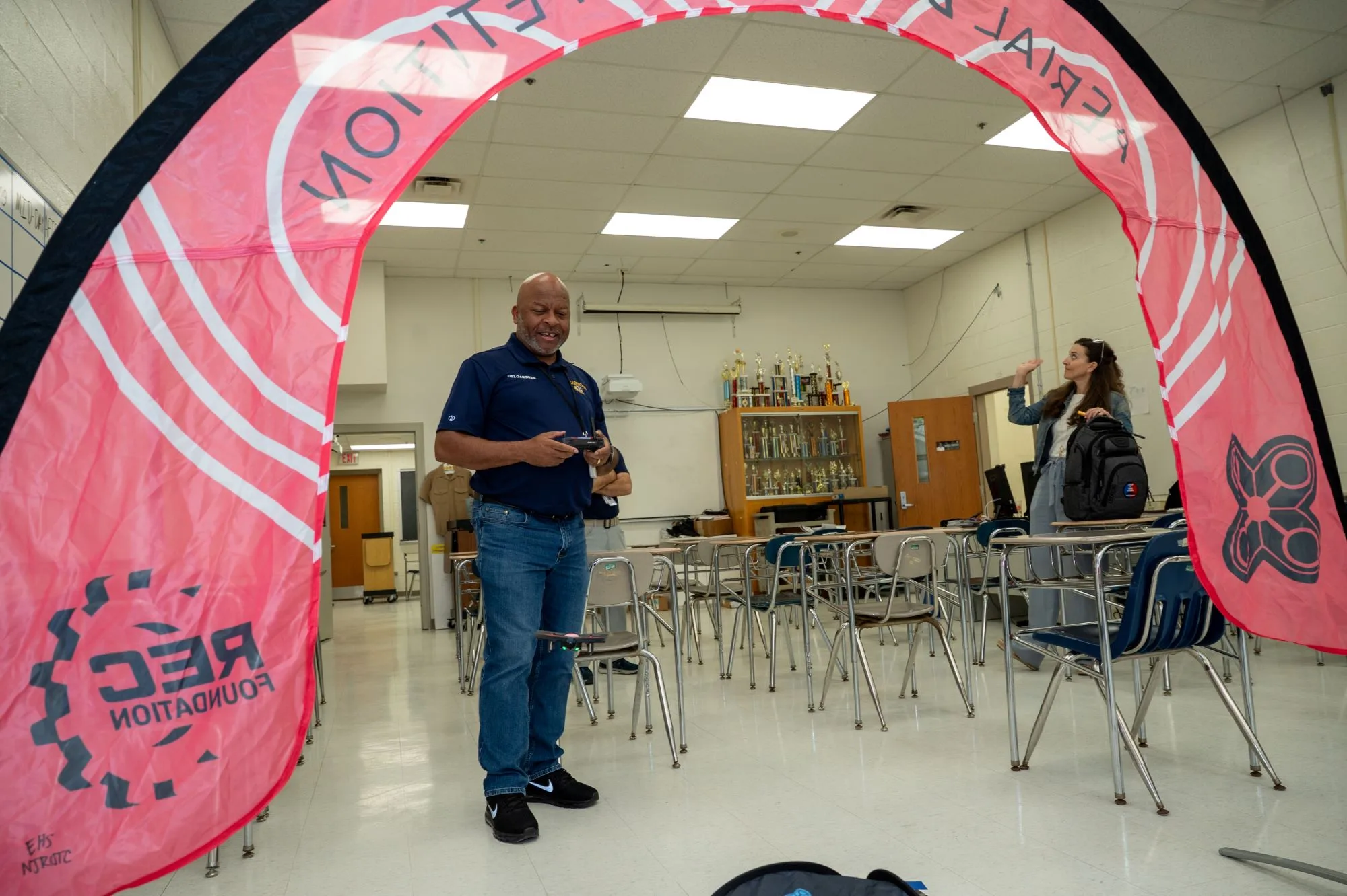 Man in a navy polo holds a drone under a pink REC Foundation arch in a classroom.
