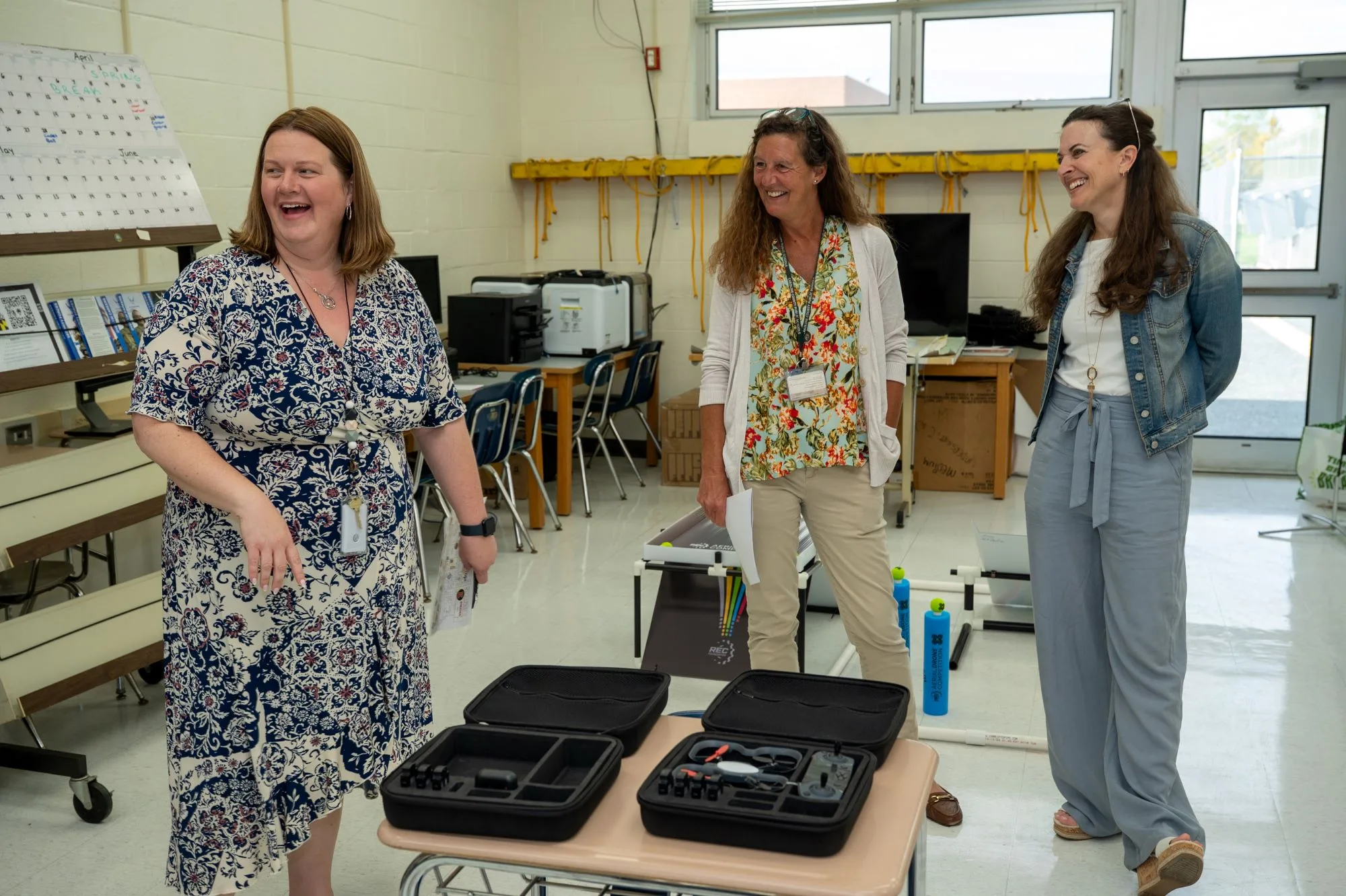 Three women laugh beside black cases holding drones on a classroom table.