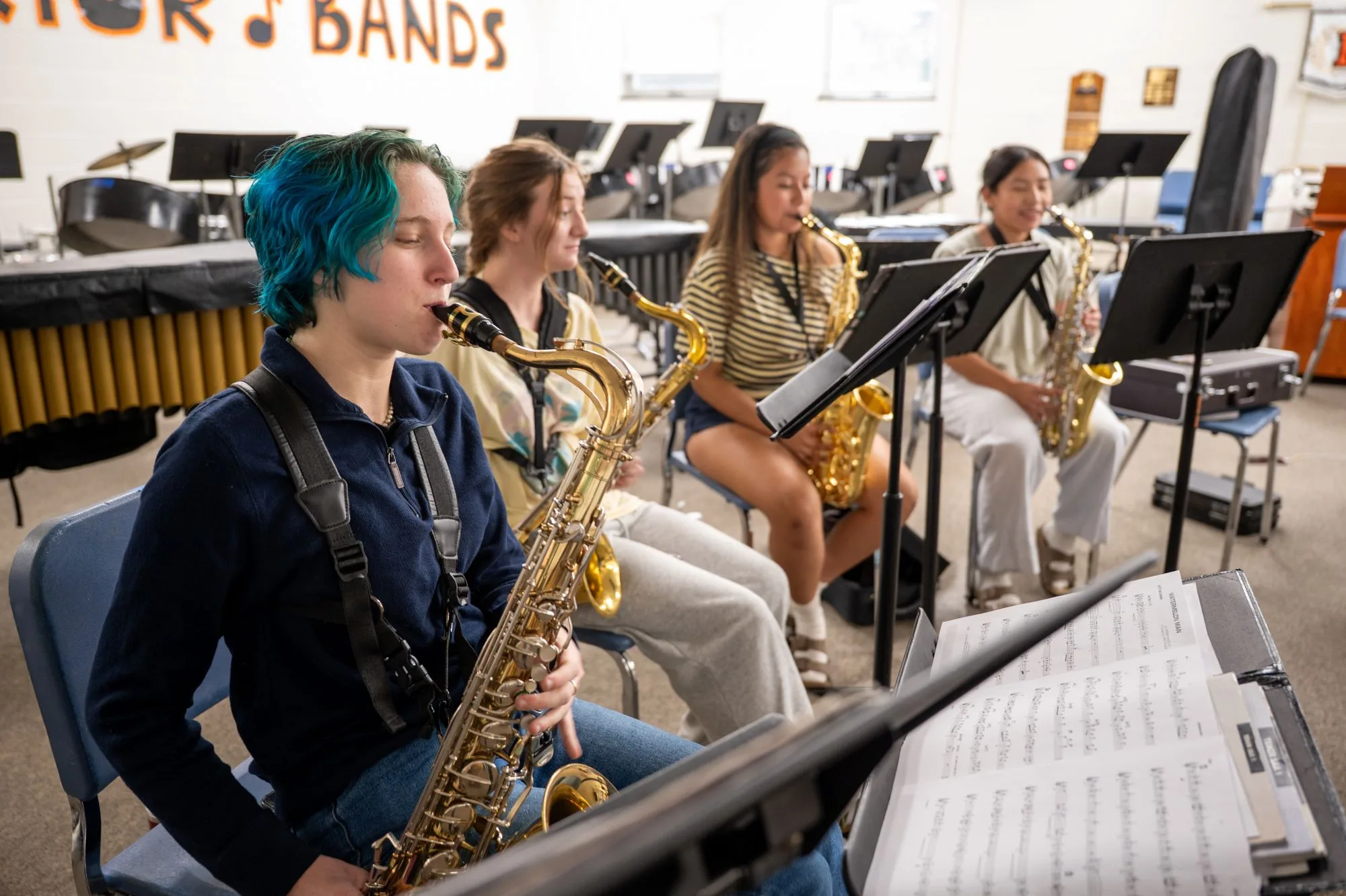 Four students play saxophones in a band room, one with teal hair in the foreground.
