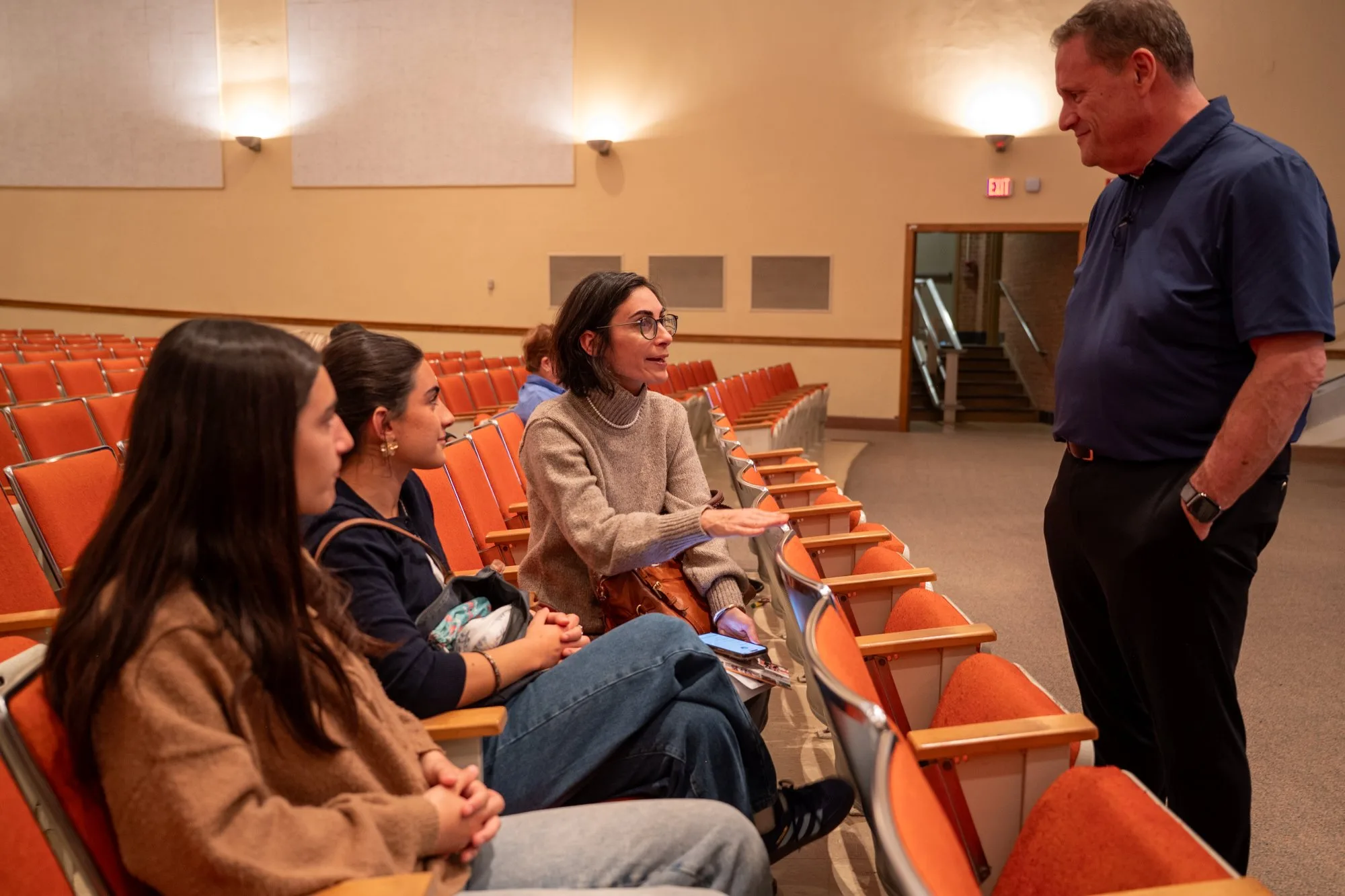 Woman in a sweater gestures to three students seated in an auditorium as a man in navy stands by.