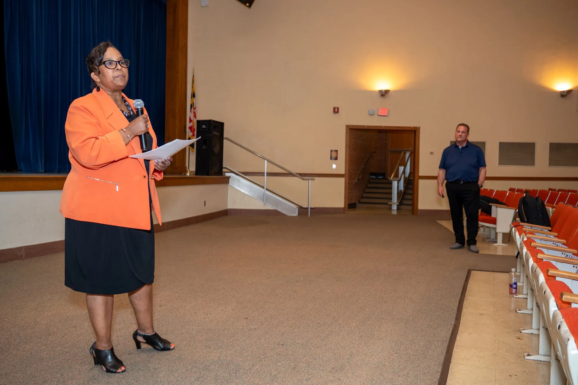 Woman in an orange blazer speaks into a microphone in an empty auditorium.