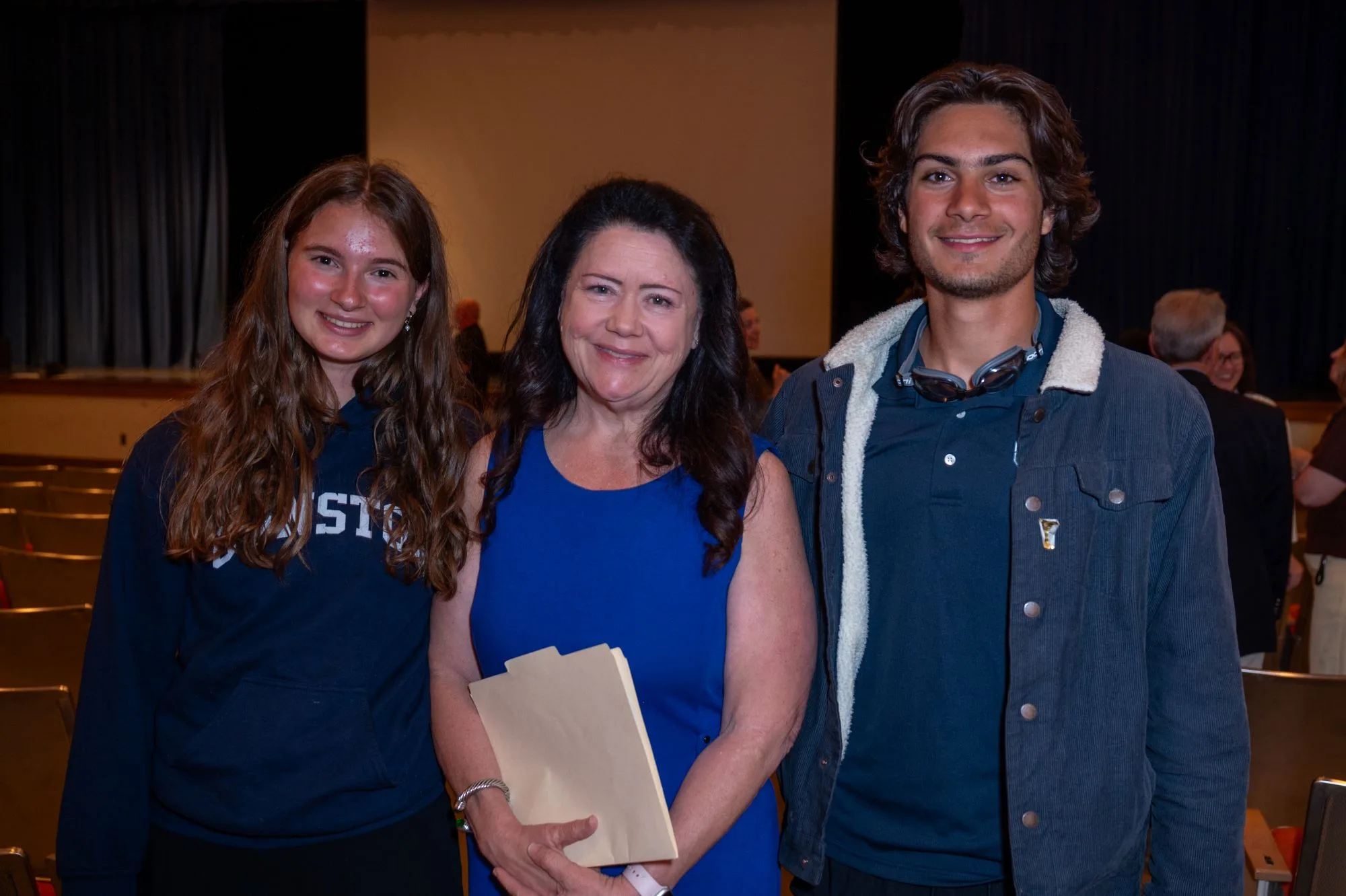 Two teens and an adult woman holding a folder smile in an auditorium.