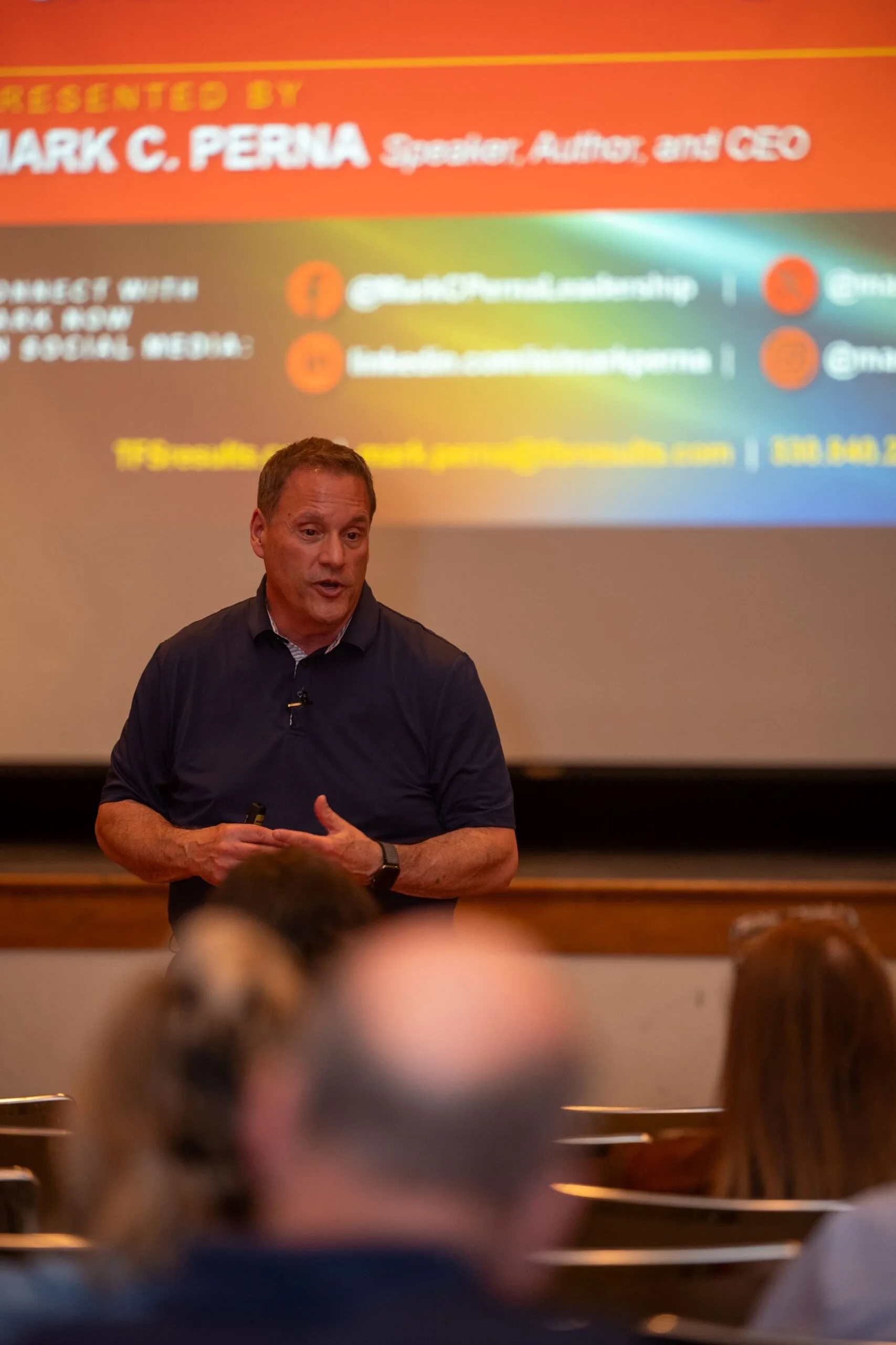 Speaker in a navy polo gestures before a slide reading Presented by Mark C. Perna, Speaker, Author, and CEO.