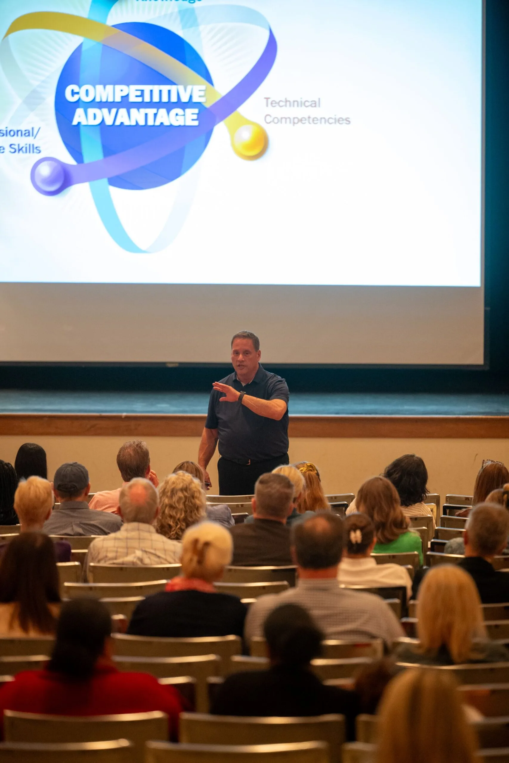 Speaker gestures before a Competitive Advantage slide while an audience sits in rows.
