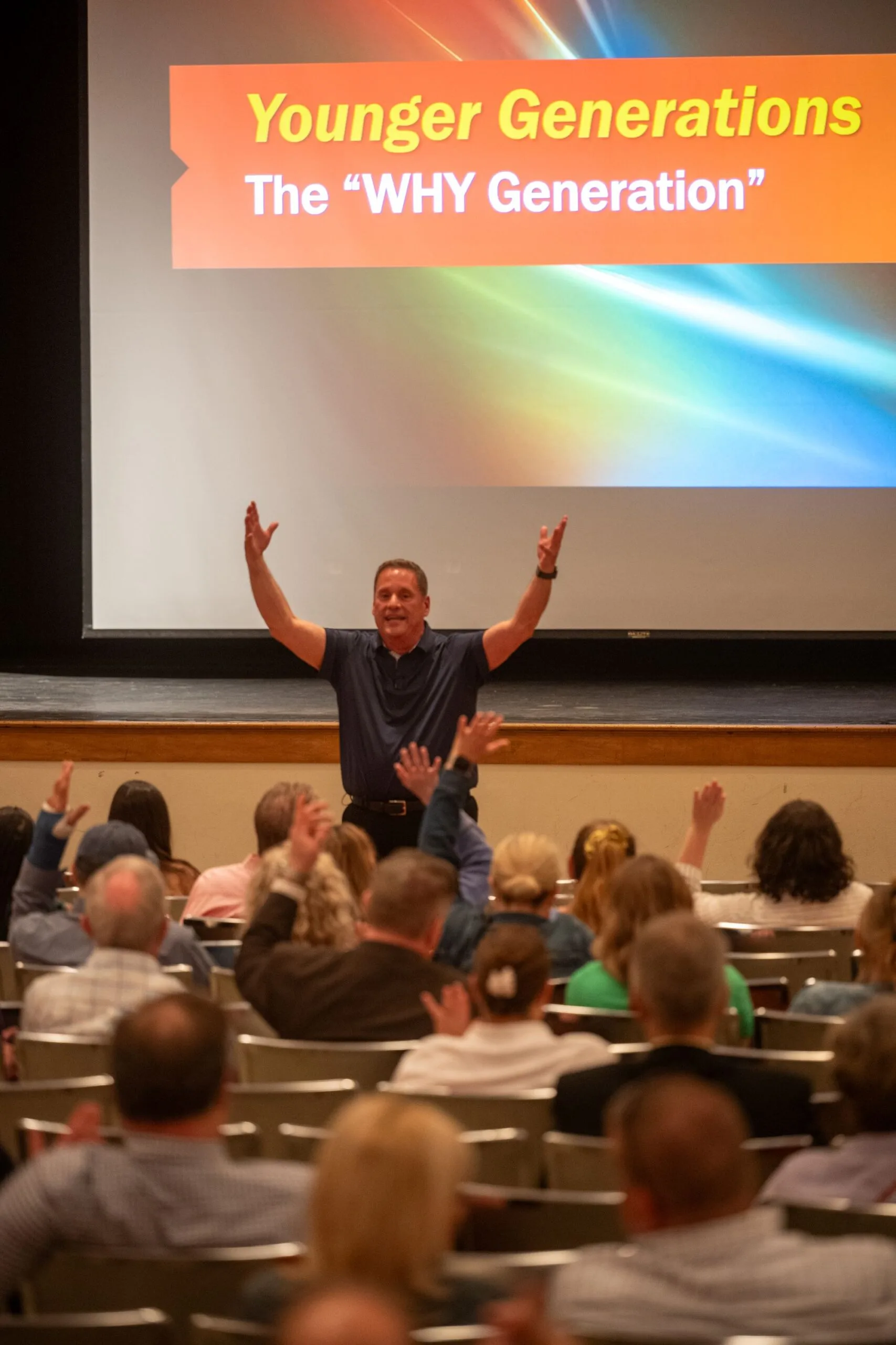 Speaker with arms raised before a slide reading Younger Generations, The WHY Generation, as hands rise in the audience.