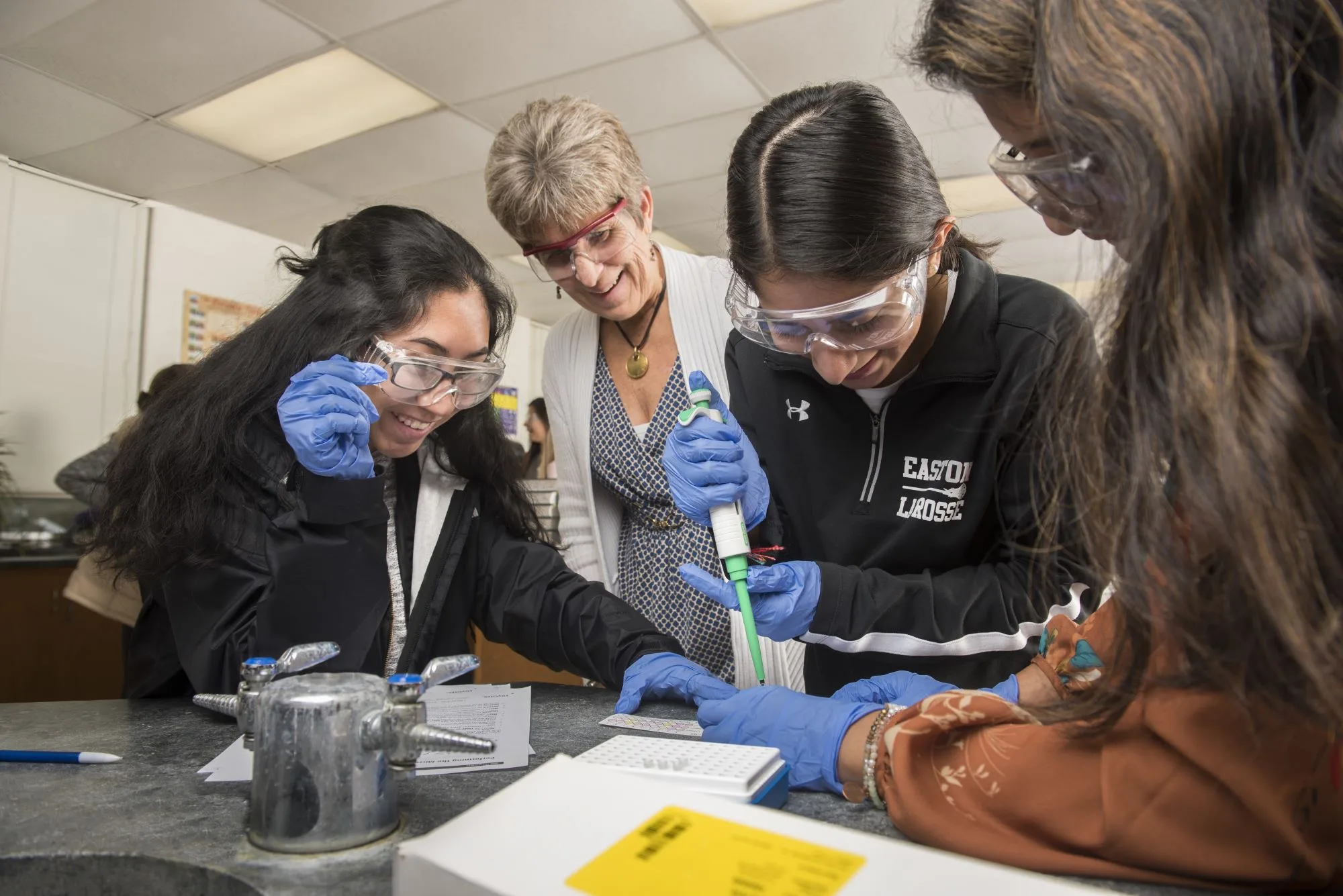 Smiling students in goggles and gloves work at a lab bench with an instructor.